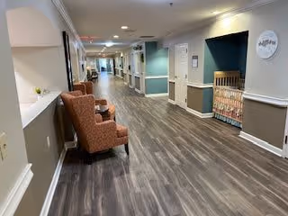 A long, well-lit hallway in a senior living facility with wood-look flooring, beige walls with white trim, and several brown upholstered chairs along the left side. There are doors and wall decorations along the right side, including a quilt hanging on the wall. The hallway appears clean and inviting.
