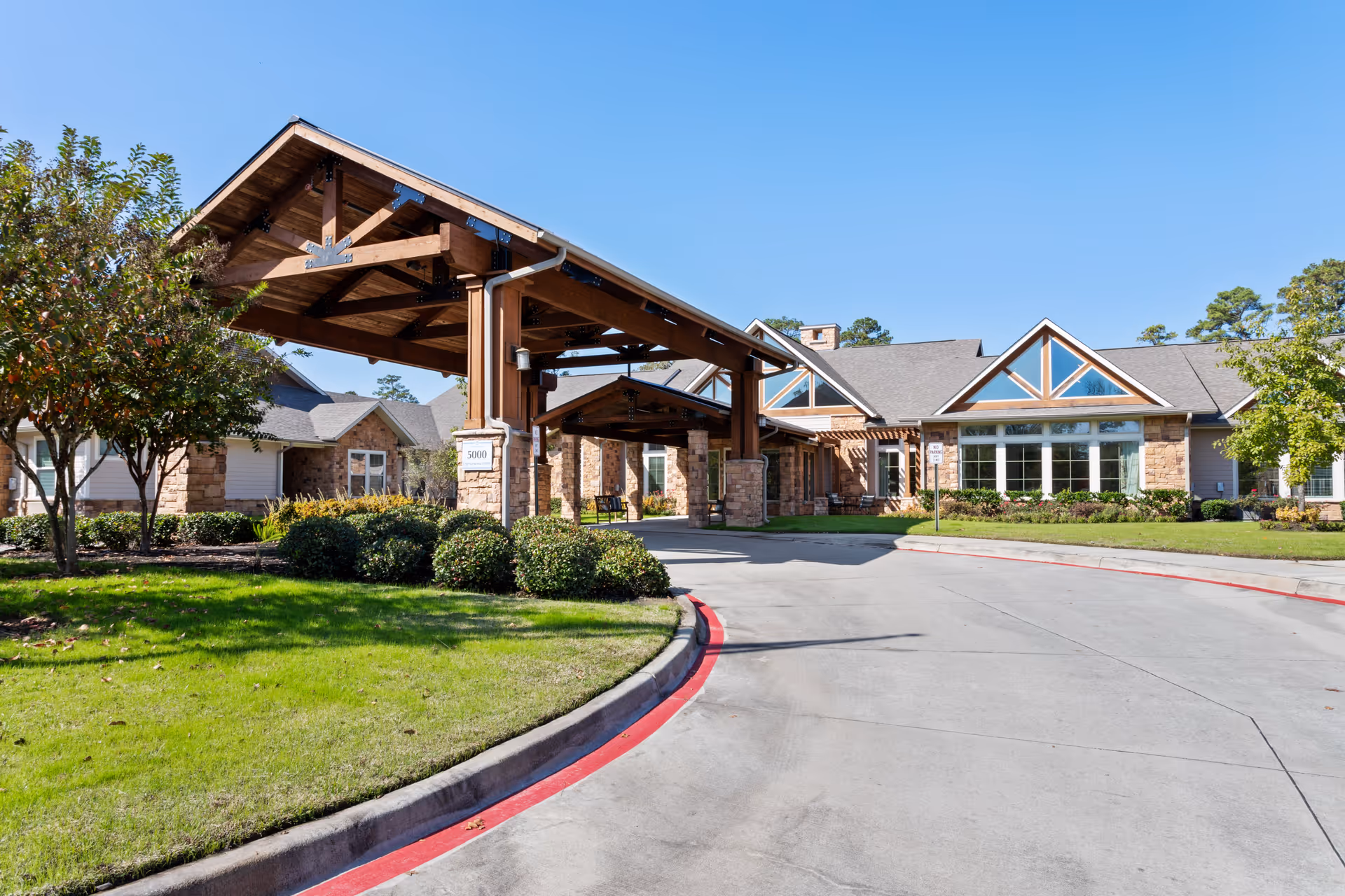 Entrance of a senior living facility with a large wooden covered driveway, stone pillars, and well-maintained landscaping including green grass, bushes, and trees under a clear blue sky.