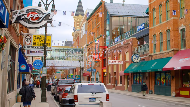A city street scene with parked cars along the curb and pedestrians walking on the sidewalk. The street is lined with brick buildings featuring various colorful signs for restaurants and shops, including a sign for Cold Stone and a neon sign for a restaurant. There are decorative flags strung across the street and a glass skywalk connecting two buildings in the background.