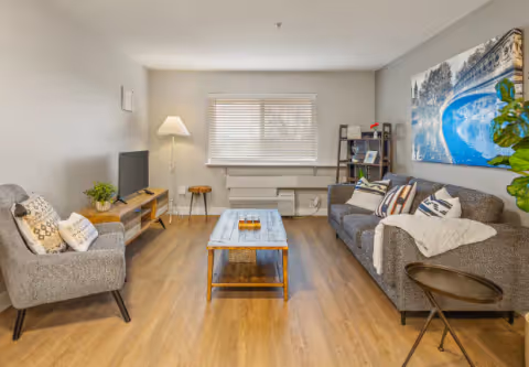 A cozy living room with a gray sofa and armchair, a wooden coffee table in the center, a TV on a wooden stand, a floor lamp, a small side table, and a large window with blinds. The room has wooden flooring and a large blue and white artwork on the wall above the sofa.