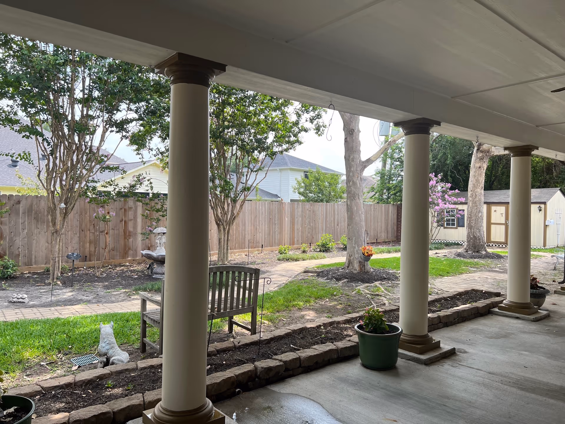 View from a covered patio with large columns looking out onto a garden area with trees, a wooden bench, a stone cat statue, a birdbath, a paved walkway, and a small shed in the background. There are potted plants on the patio and a wooden fence enclosing the garden.