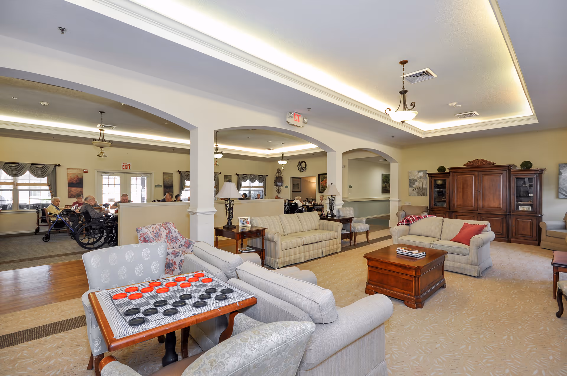 A spacious senior living facility common area with comfortable sofas, armchairs, and wooden coffee tables. A checkers game is set up on a small table in the foreground. In the background, several elderly residents are seated, some in wheelchairs, near large windows with curtains. The room has soft lighting with ceiling fixtures and a large wooden cabinet against the wall.