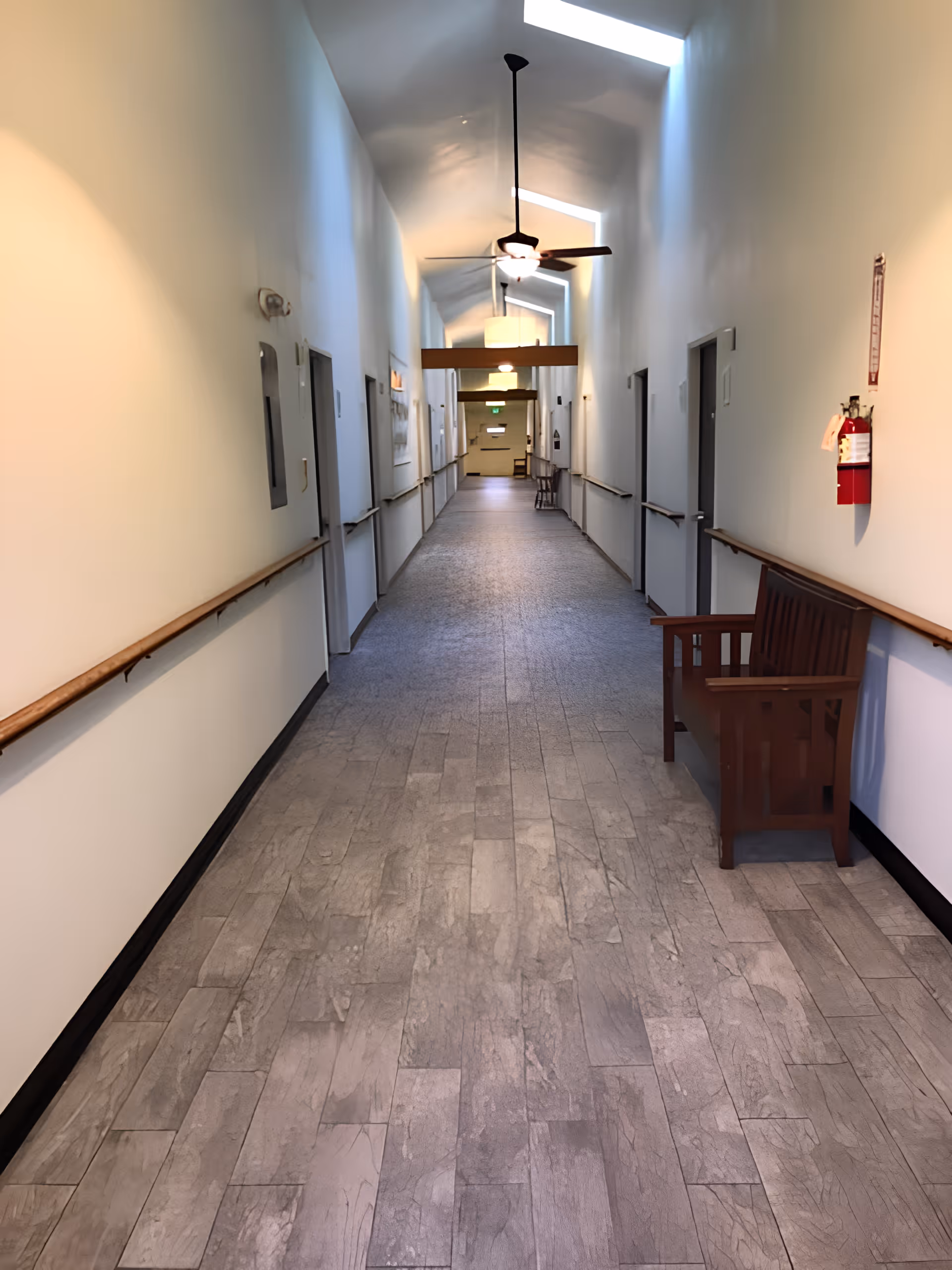 Long, well-lit interior corridor with handrails, skylights, ceiling fans, doors along both sides, and a wooden bench.