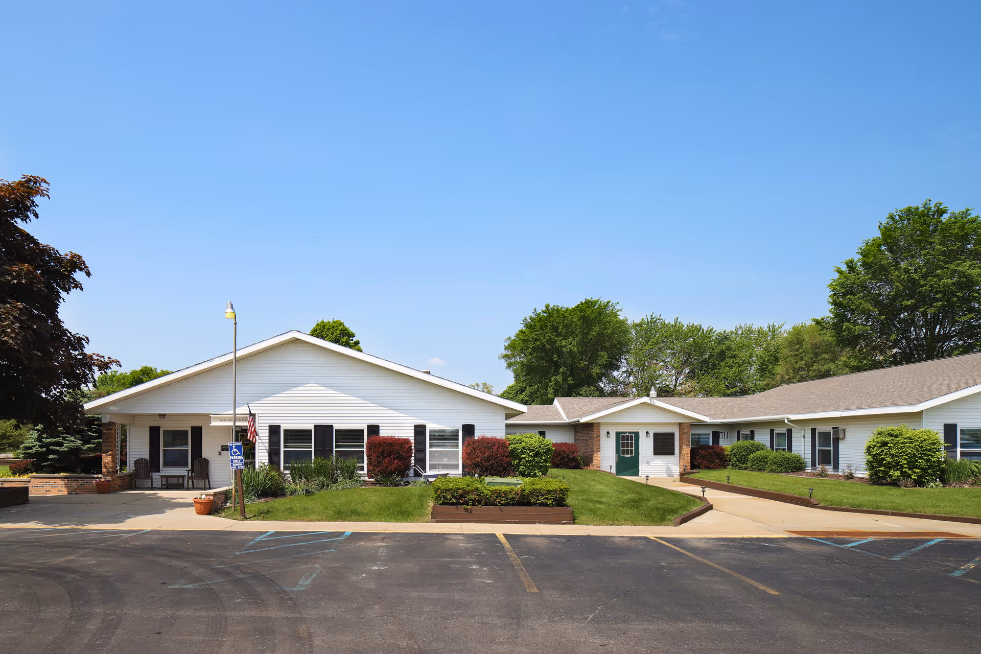 Exterior view of Divine Living Center of Mt Pleasant, a single-story white building with a sloped roof, surrounded by green shrubs and trees under a clear blue sky. There is a paved parking area in front with marked handicap parking spaces and a small ramp leading to the entrance.