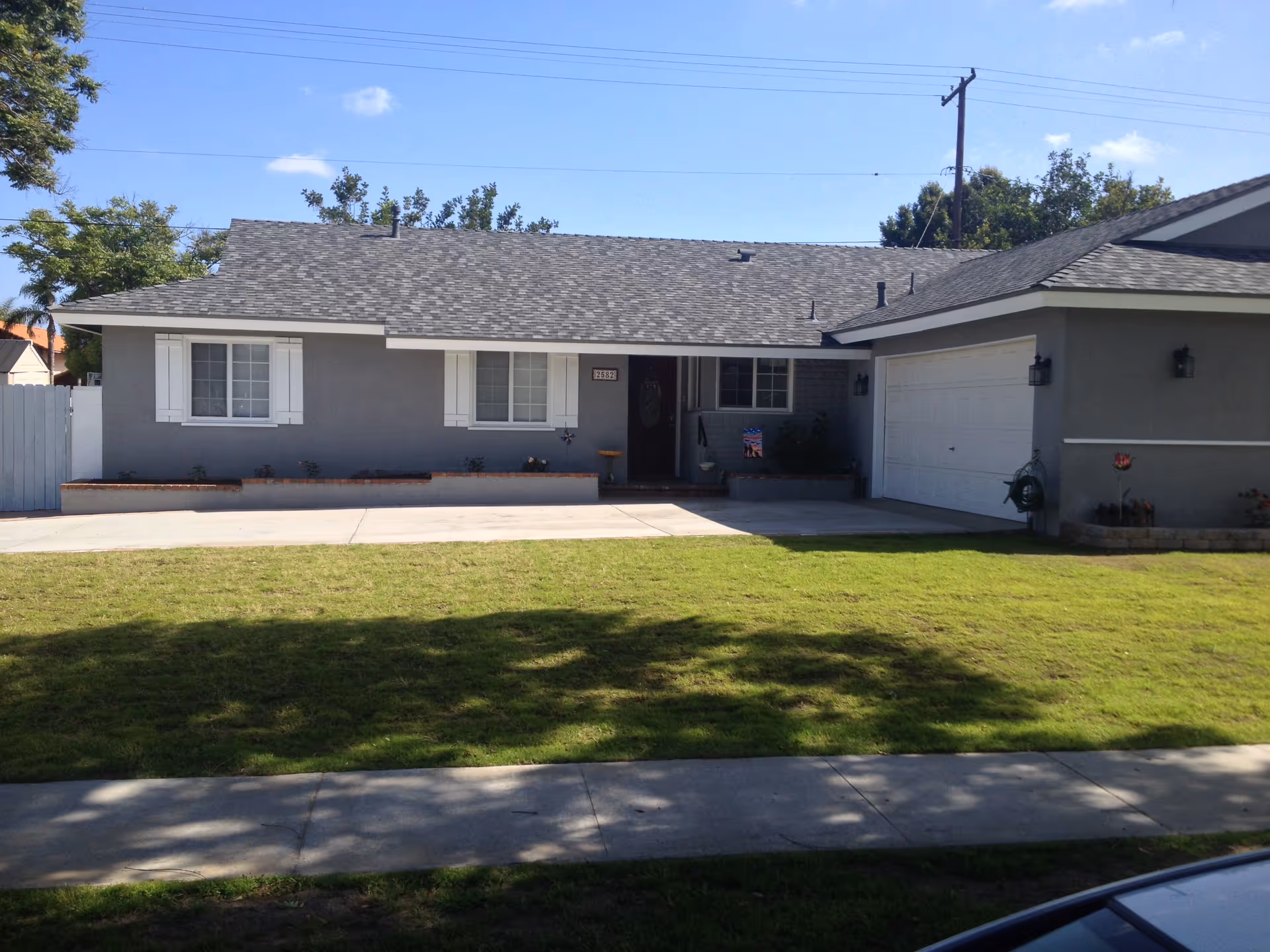 Front exterior view of a single-story gray house with a shingled roof, two white-framed windows with shutters, a front door with a small porch, a two-car garage, and a well-maintained lawn with a sidewalk in front.