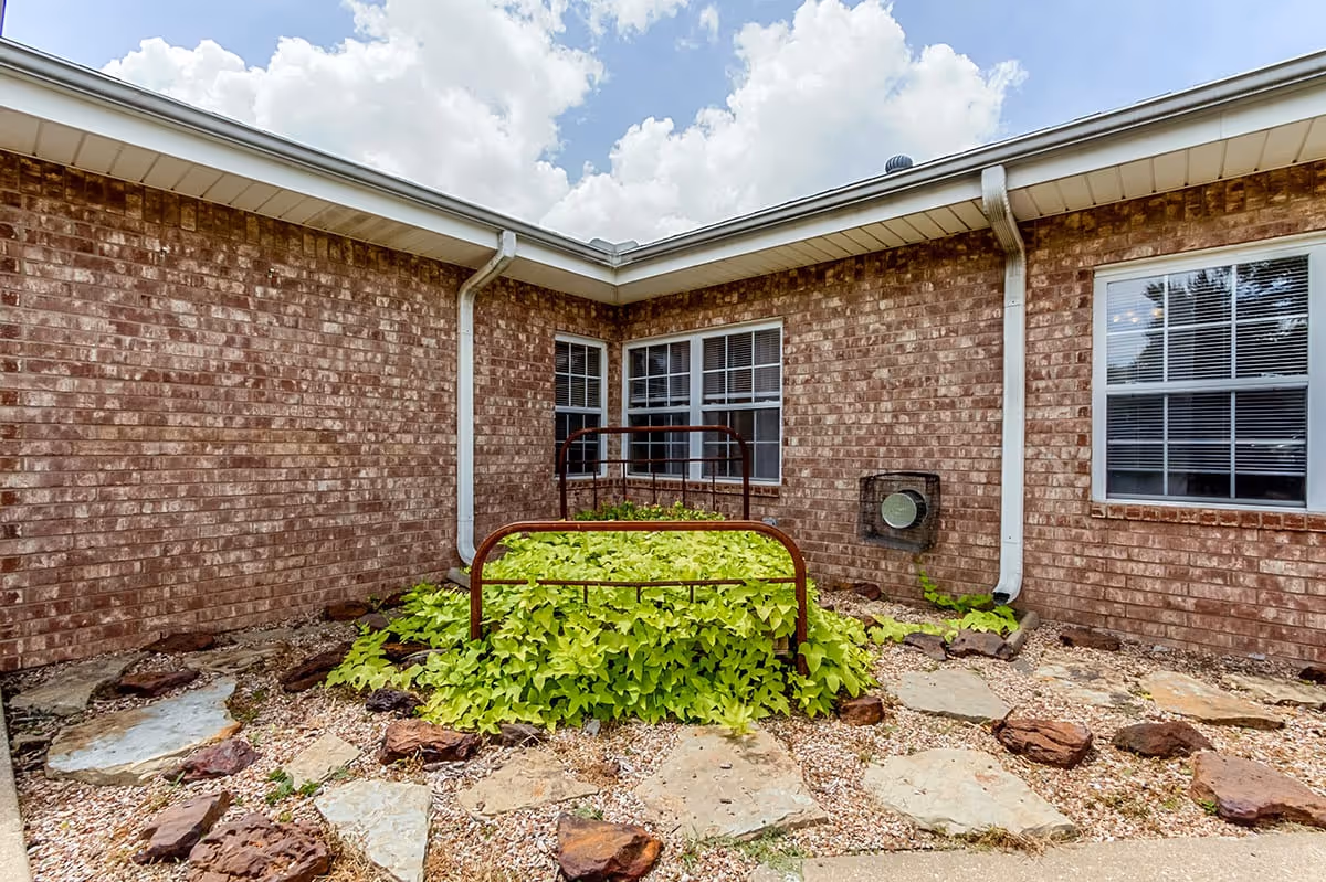 Brick courtyard corner with a decorative metal bed frame filled with green plants, stone pathway and windows.