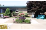 Outdoor patio area at St Anne Home featuring a central garden bed with flowers and plants, surrounded by concrete walkways. There are several benches and chairs in white, yellow, and turquoise colors placed around the patio. A large tree provides shade on the right side, and the building with windows is visible in the background.