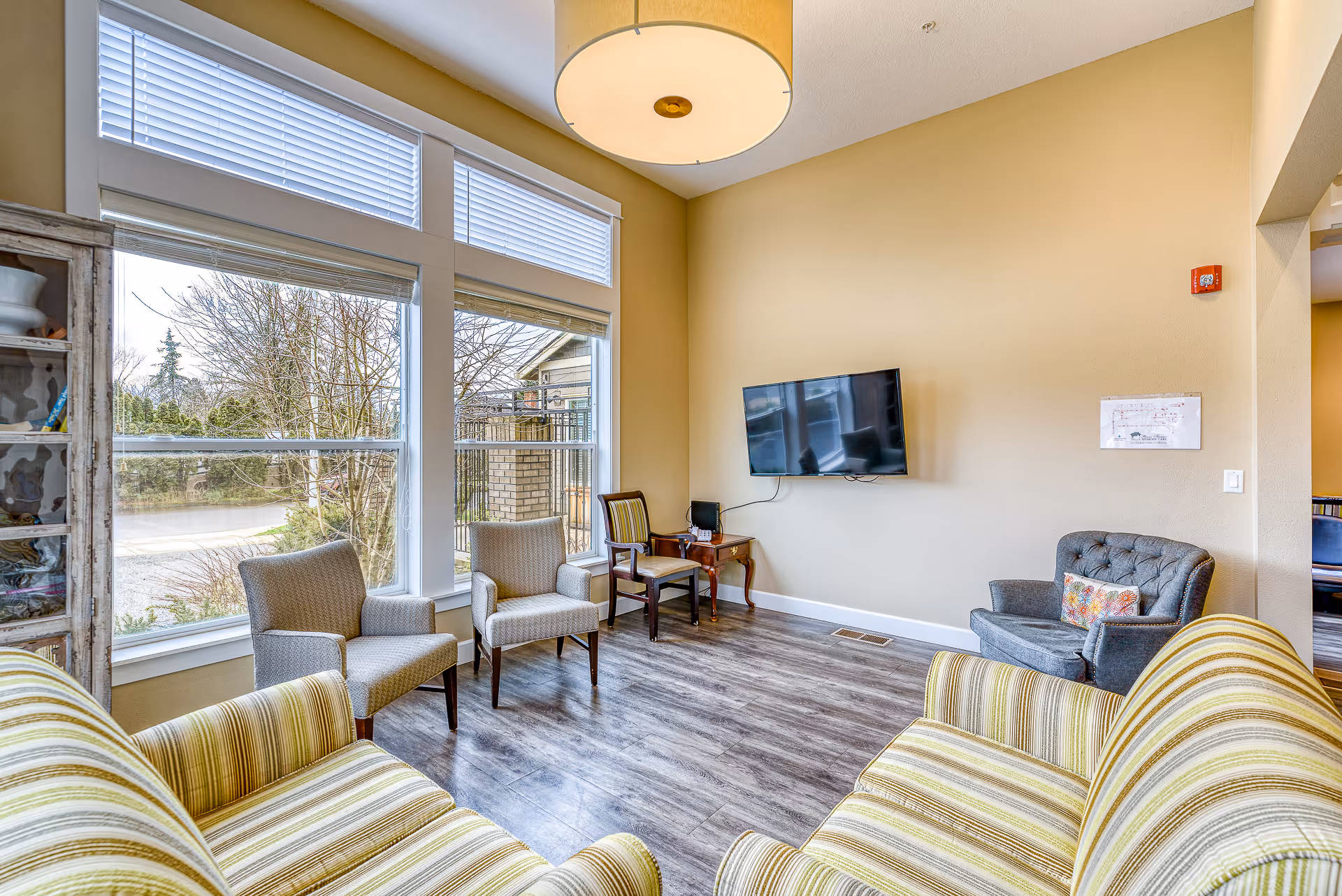 Bright communal living room with striped sofas, armchairs, large windows and a wall-mounted TV.