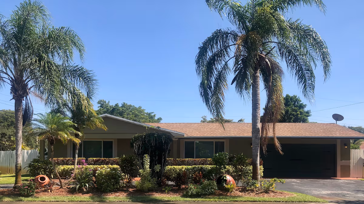 Single-story residential building with a brown roof and beige exterior, surrounded by palm trees and well-maintained landscaping under a clear blue sky.