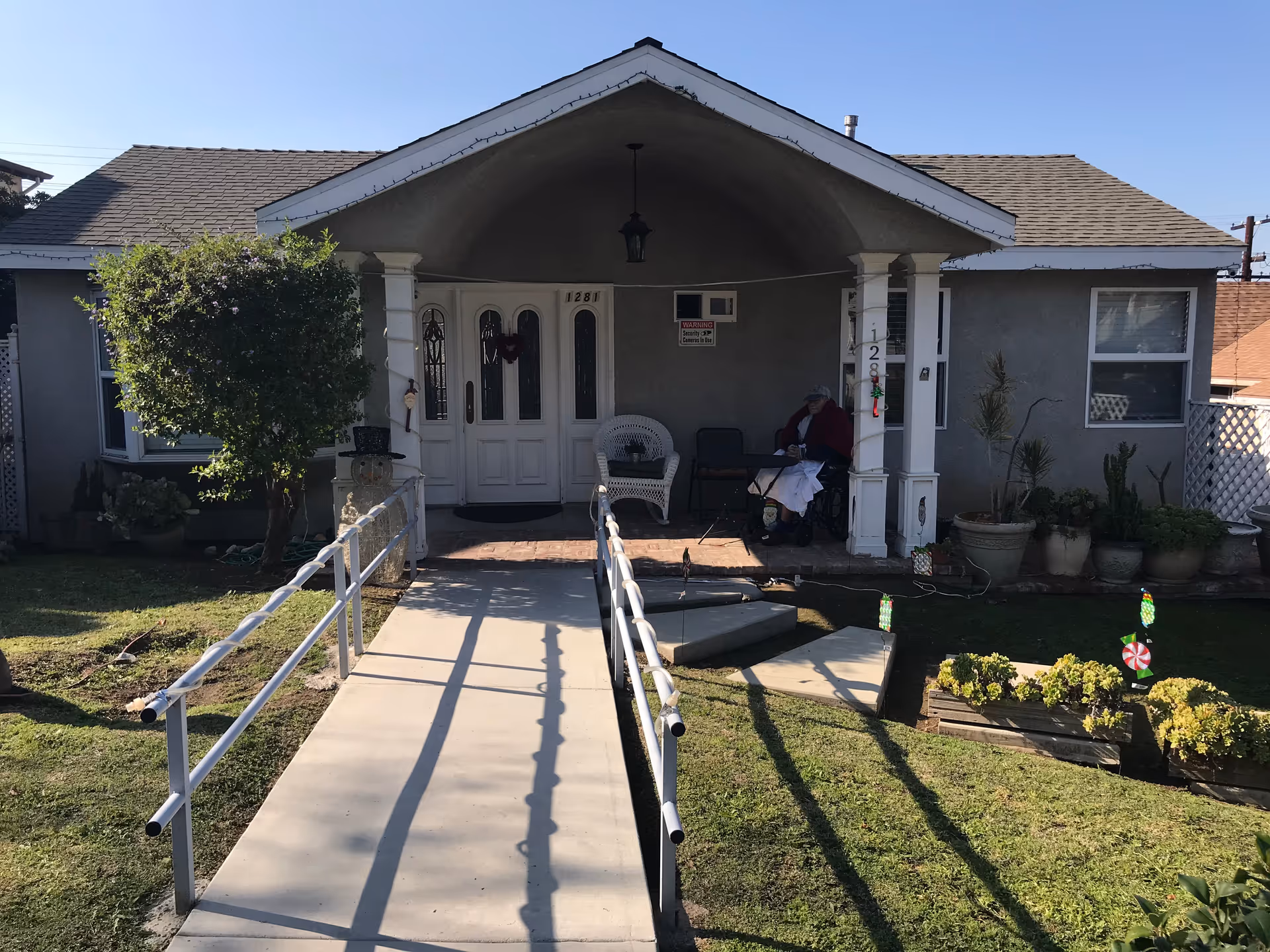 Single-story house front with a concrete wheelchair ramp leading to a covered porch where a person sits beside potted plants.