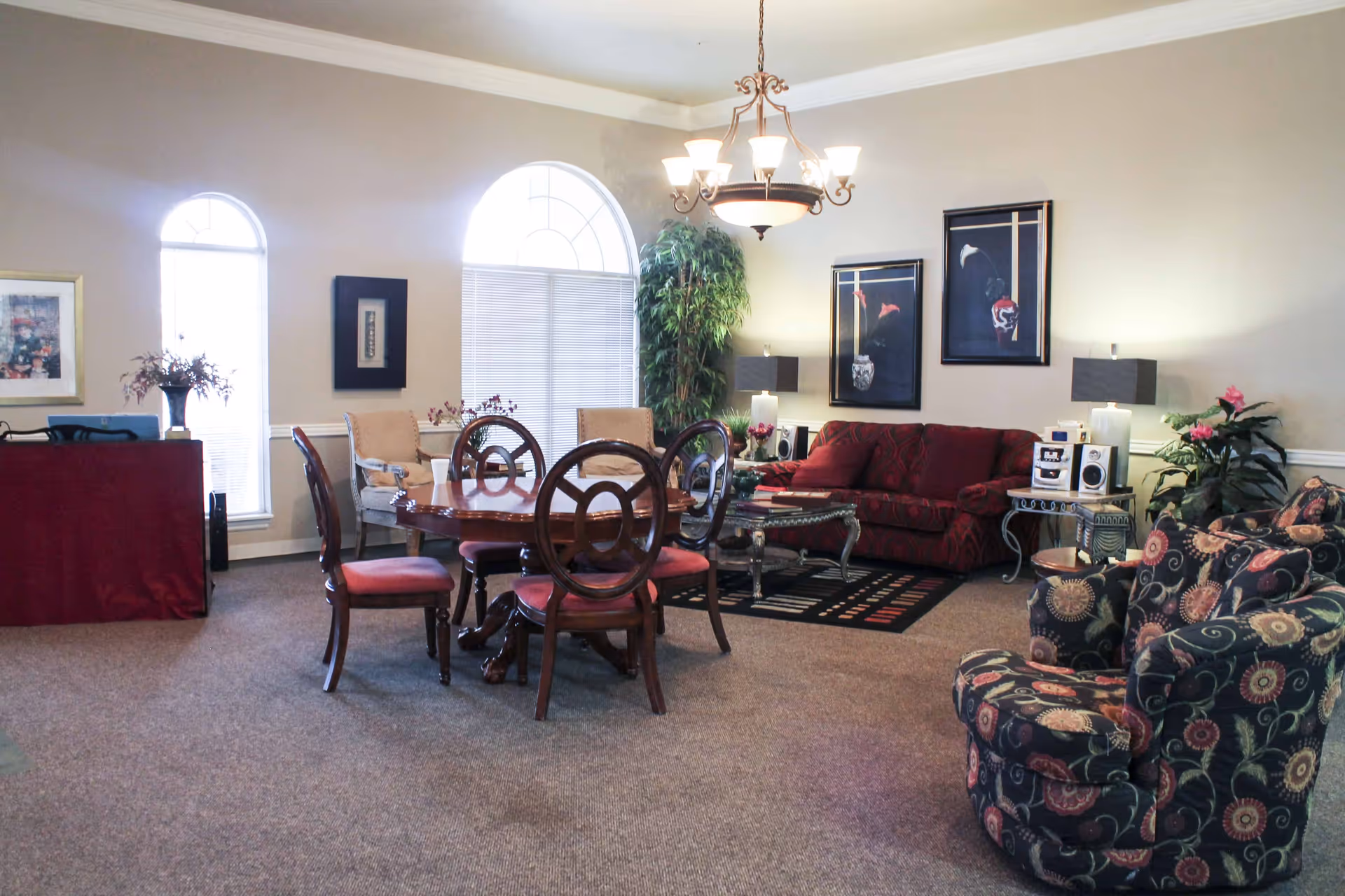 A spacious living room with a round wooden dining table surrounded by four chairs with red cushions. There is a red sofa and a patterned armchair, along with two side tables holding lamps and a stereo system. The room has large arched windows with blinds, two framed artworks on the wall, a chandelier hanging from the ceiling, and several potted plants adding greenery.