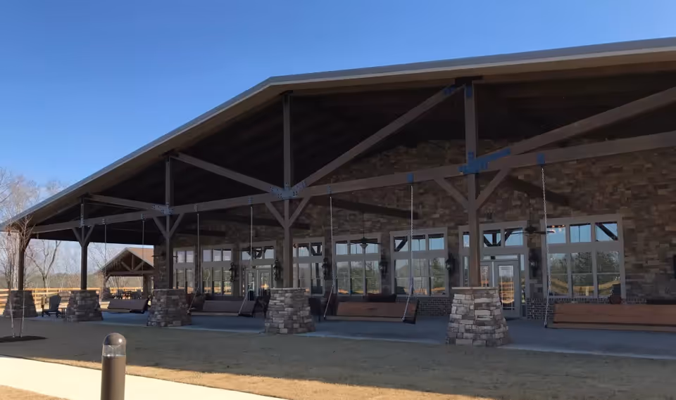Front exterior of a large building with a wide covered porch supported by timber beams and stone pillars, featuring hanging wooden swings and large windows.