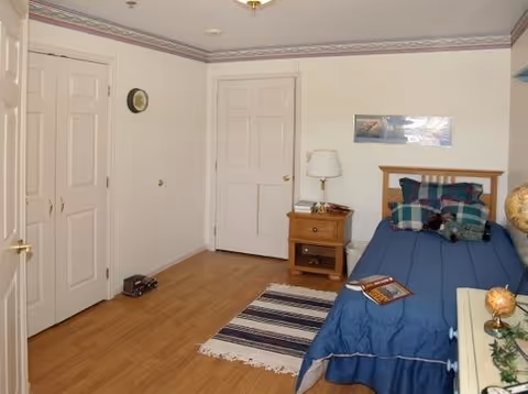 A simple bedroom with a twin bed dressed in blue, a nightstand with a lamp, hardwood floors, and closet doors.