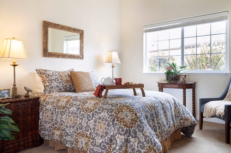 A cozy bedroom with a patterned bedspread on a bed, two bedside tables each with a lamp, a decorative mirror on the wall above the bed, a window letting in natural light, a small wooden table with plants, and a chair with a throw blanket.