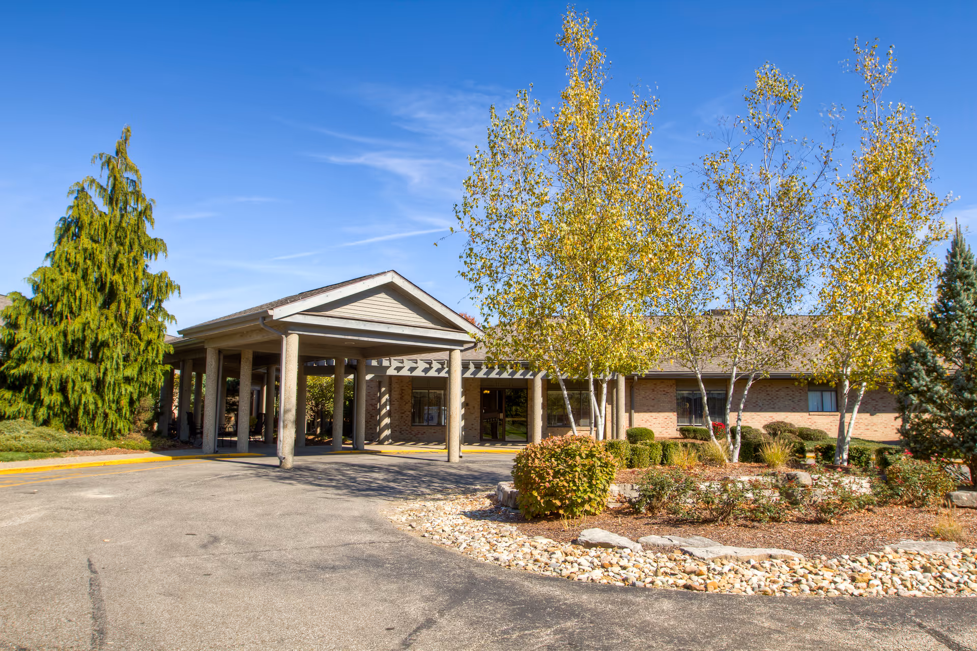 Exterior view of Brookdale Zanesville facility showing the entrance with a covered driveway, surrounded by landscaped bushes, trees with yellow leaves, and a clear blue sky.