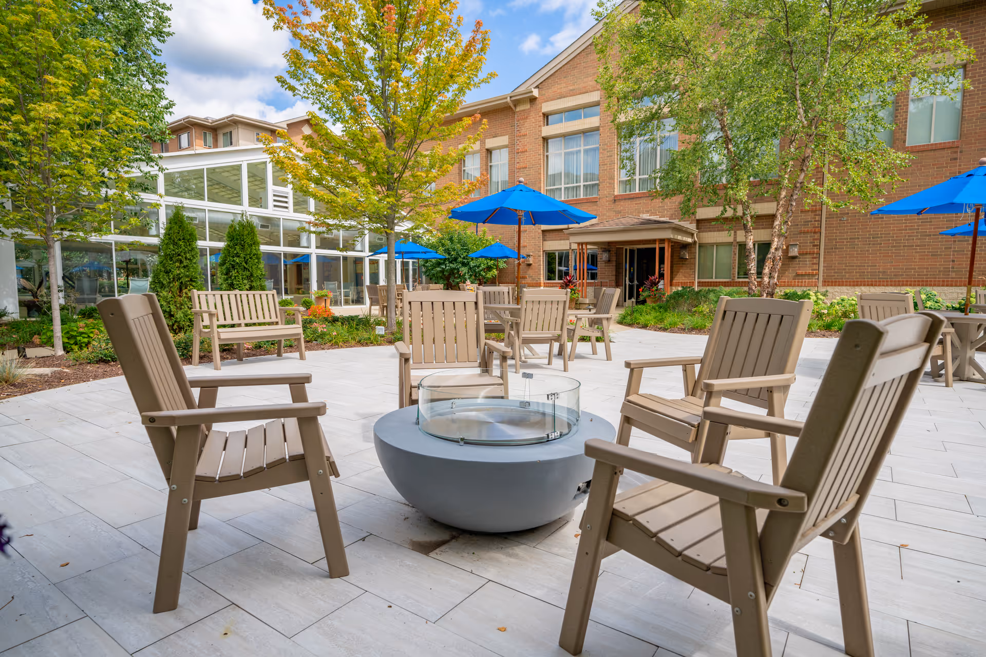 Outdoor patio area at Monarch Landing with beige wooden chairs arranged around a circular fire pit. There are additional tables with blue umbrellas, green trees, and a brick building in the background under a partly cloudy sky.