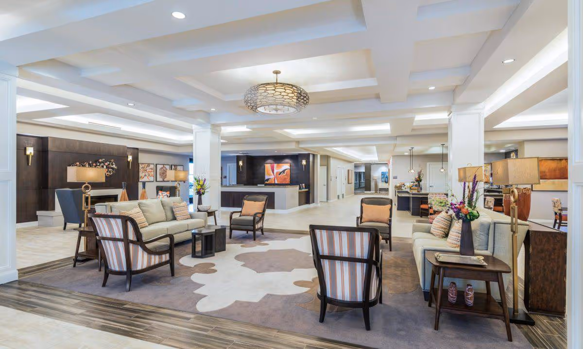 Bright, spacious senior living lobby with sofas, armchairs, side tables, decorative lighting and a reception desk in the background.
