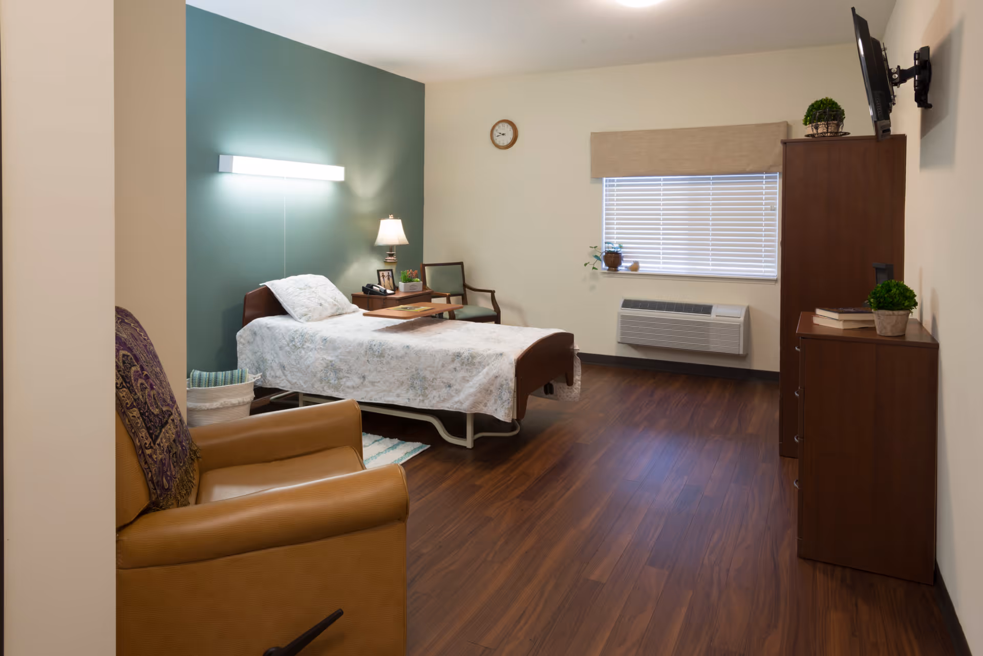 A neatly arranged bedroom in a senior living facility featuring a single bed with a floral bedspread, a wooden nightstand with a lamp and small plants, a comfortable armchair with a patterned throw, a wooden dresser with books and a potted plant, a wall-mounted TV, a window with blinds, and a wall clock.