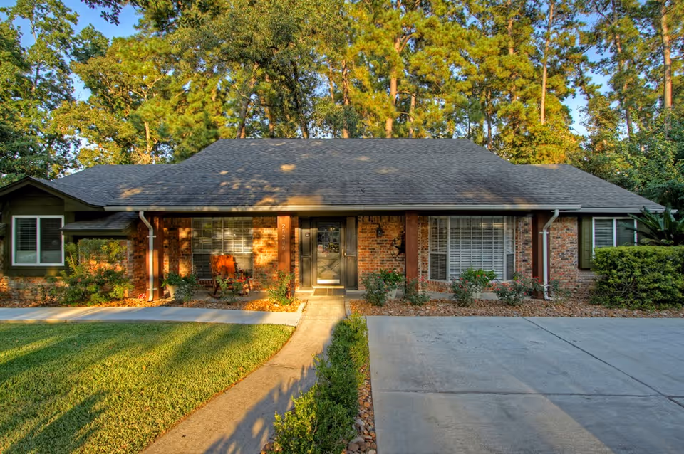 Front exterior view of a single-story brick house with a dark shingled roof, large windows with white blinds, a concrete driveway, and a walkway leading to a black front door. The house is surrounded by green grass, shrubs, and tall trees in the background.
