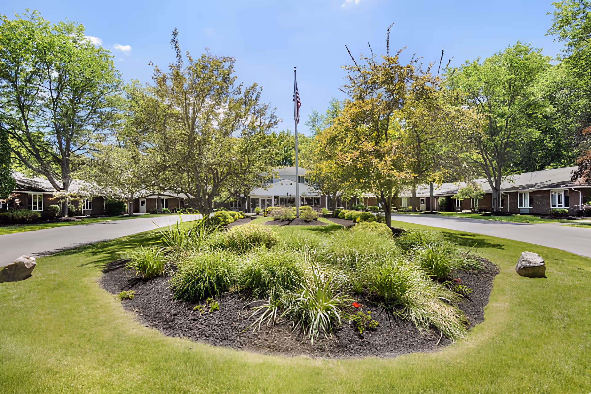 A landscaped garden area with green grass, bushes, and trees surrounding a flagpole with an American flag in the center. The garden is bordered by a paved driveway leading to a single-story building with multiple windows, under a clear blue sky.