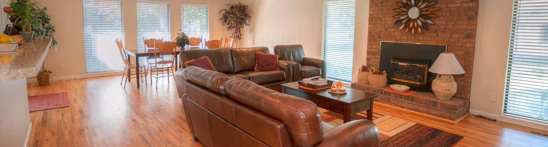 Open-plan living area with brown leather sofas facing a brick fireplace and a dining table by large windows.