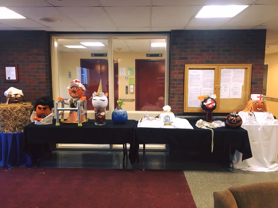 A row of decorated pumpkins and seasonal crafts displayed on tables in a senior living facility lobby hallway.