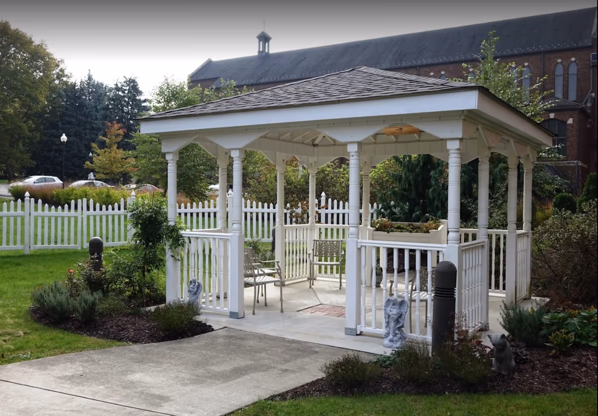 A white wooden gazebo with chairs in a landscaped garden in front of a white picket fence and a large brick building.