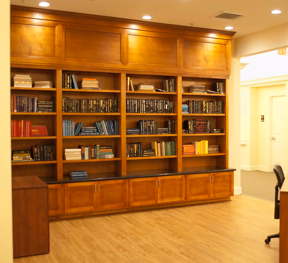 Interior view of a room with a large wooden bookshelf filled with books of various colors and sizes. The bookshelf has cabinets at the bottom and is illuminated by ceiling lights. The room has light-colored wooden flooring and light-colored walls. There is a doorway leading to another room on the right side.