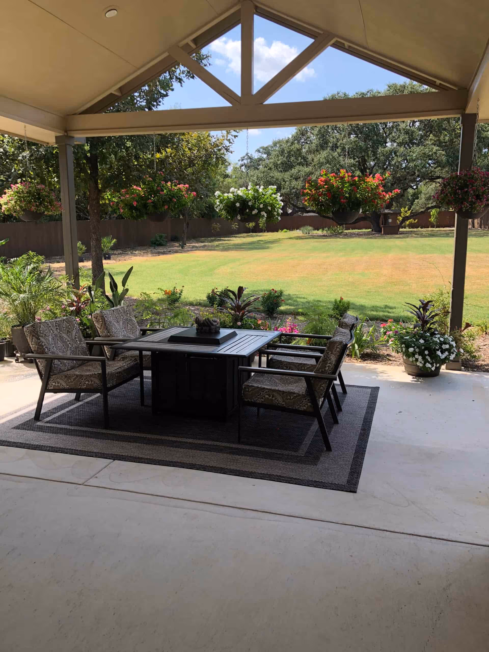 Covered outdoor patio area with a set of four cushioned chairs around a square table on a large rug. Hanging flower baskets and potted plants surround the patio, overlooking a spacious green lawn with trees and a wooden fence in the background under a partly cloudy sky.