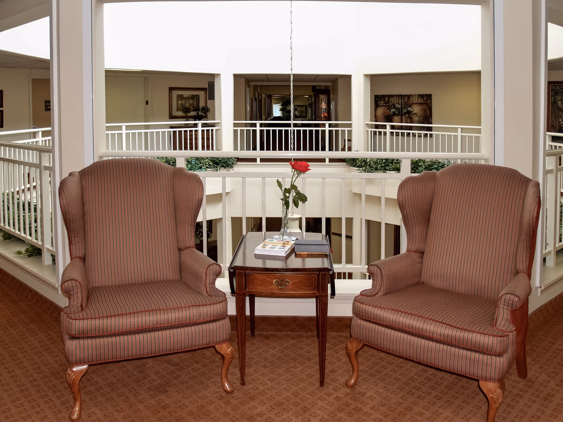 Two striped upholstered armchairs with wooden legs positioned on either side of a small wooden table with a drawer. On the table, there is a glass vase holding a single red rose and a few books. The seating area overlooks an interior atrium with white railings and a hallway in the background.