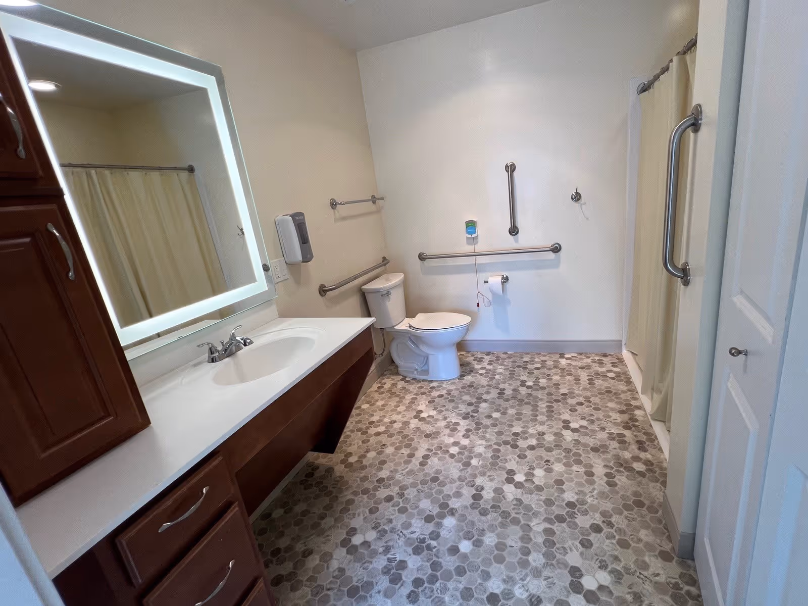 Accessible bathroom with a sink and illuminated mirror, toilet with multiple grab bars, and a shower with a curtain.