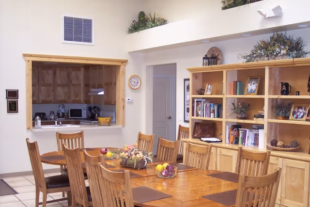 Dining room with a large wooden table and chairs, a serving counter to the kitchen, and wooden shelving with books and decorations.