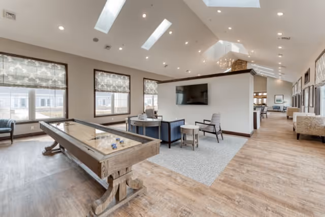 Bright communal lounge with a shuffleboard table, seating area, large windows, skylights, and a wall-mounted TV.