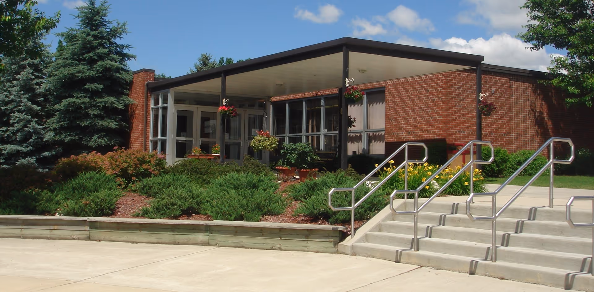 Exterior view of a single-story brick building with large windows and a covered entrance. There are concrete steps with metal railings leading up to the entrance, surrounded by well-maintained green shrubs, flowering plants, and tall evergreen trees under a partly cloudy sky.