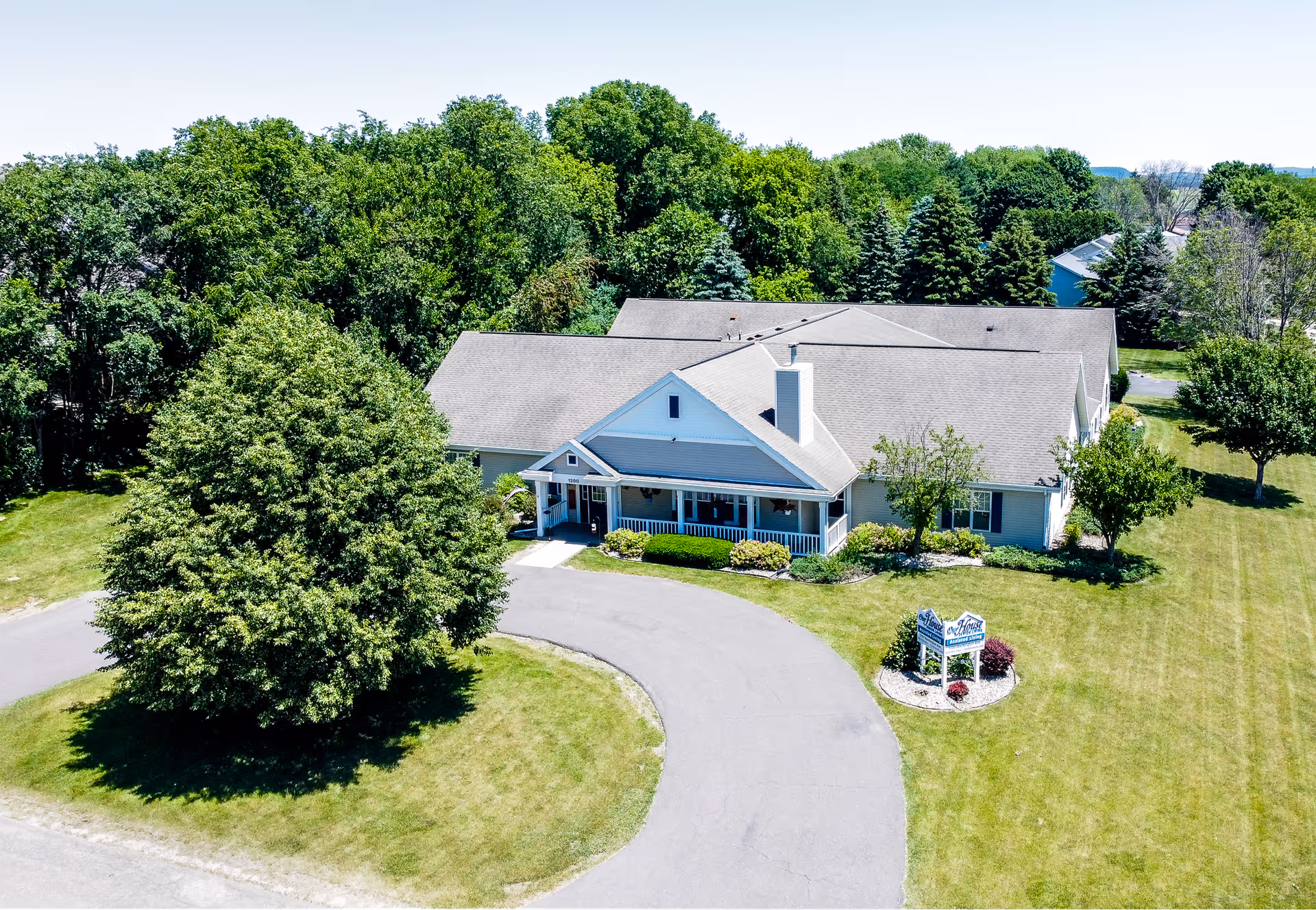 Aerial view of a single-story assisted living building with a circular driveway, front porch, lawn, and surrounding trees.