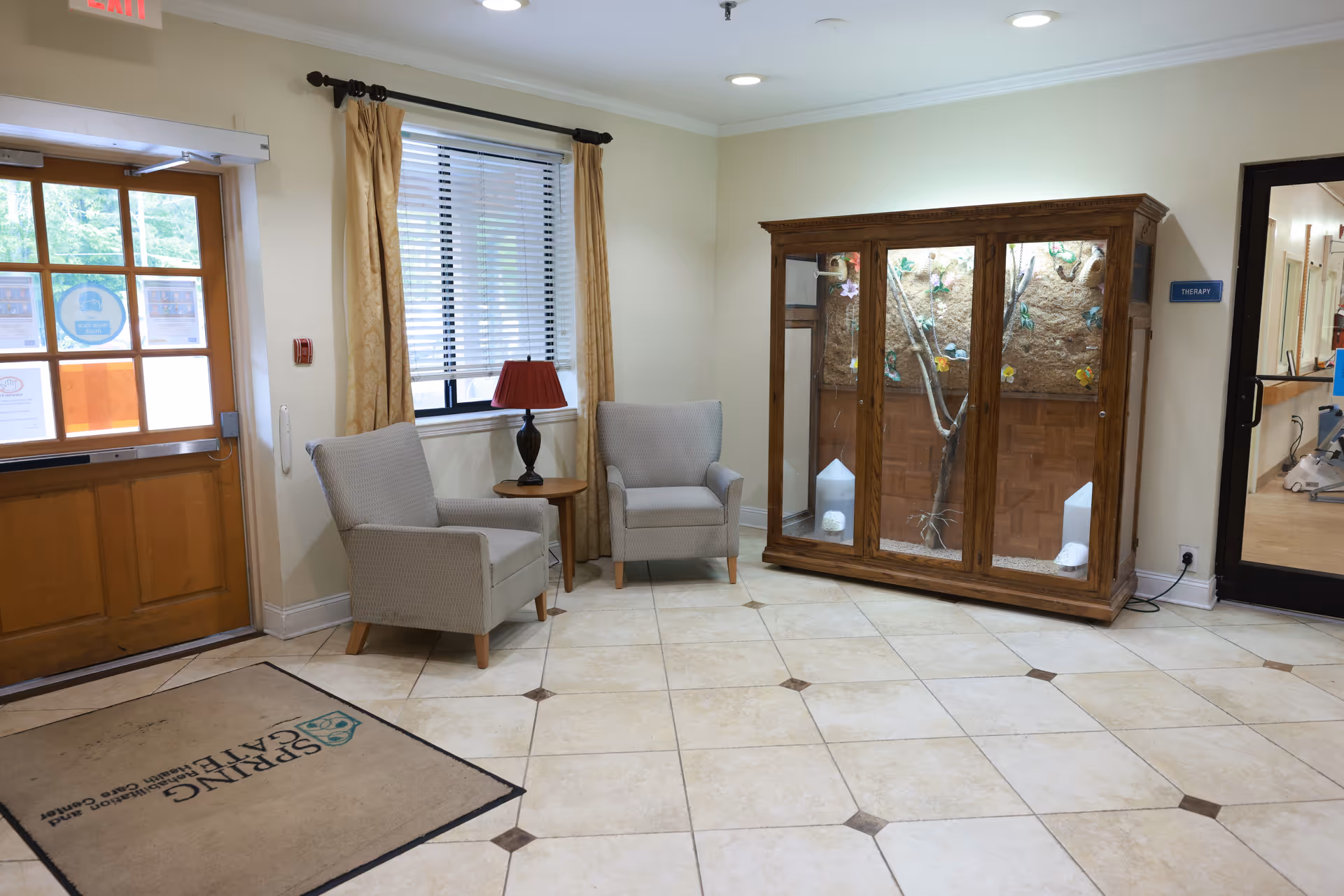 Small lobby interior with two armchairs and a side table by a window, a wooden display case, and an entrance door.
