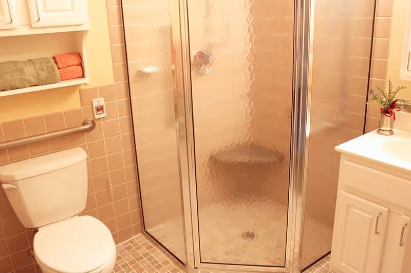 A bathroom with a white toilet, a grab bar on the wall, a glass-enclosed shower with textured glass doors, a white vanity with a sink, and a small vase with flowers on the countertop. There are shelves above the toilet holding folded towels.