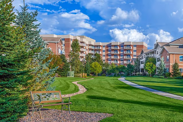 A well-maintained outdoor garden area with a green lawn, a winding stone path, a wooden bench, and various trees. In the background, there are multi-story residential buildings under a partly cloudy blue sky.