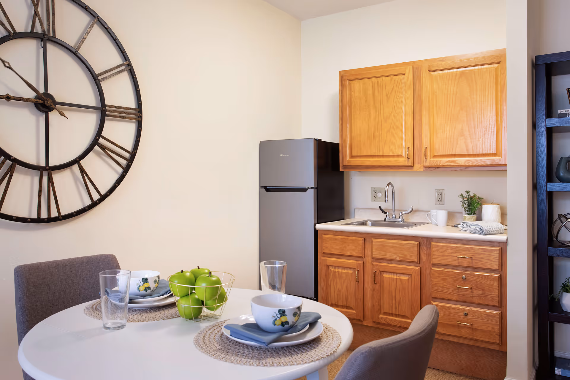 A small kitchen area with wooden cabinets, a stainless steel refrigerator, and a sink. In front of the kitchen is a round white dining table set with two place settings, each with a bowl, plate, napkin, and glass. A wire basket filled with green apples is on the table. A large decorative wall clock is mounted on the wall behind the table.
