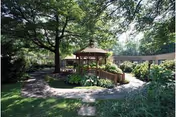 A peaceful outdoor garden area with a wooden gazebo surrounded by lush green trees, bushes, and plants. A paved walkway leads to the gazebo, and a building is visible in the background.