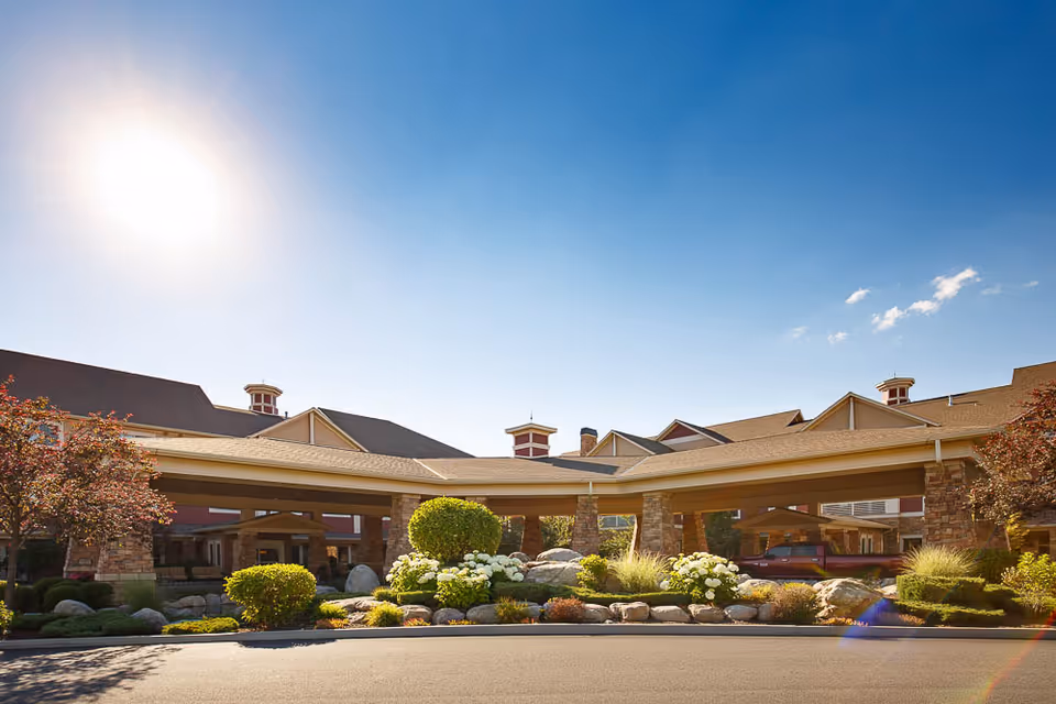 Exterior view of Garden Plaza Of Post Falls senior living facility under a clear blue sky with the sun shining brightly. The building features a covered entrance with stone pillars, landscaped bushes, flowers, and rocks in front.