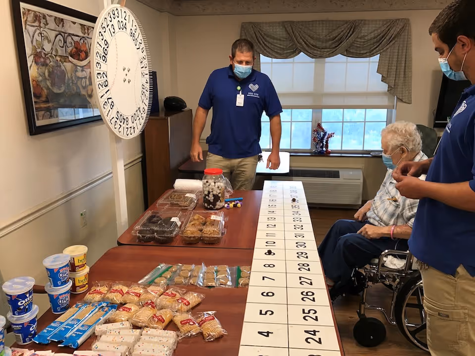 Staff members and an elderly resident wearing masks stand around a long table covered with numbered cards and snacks in a nursing facility room.