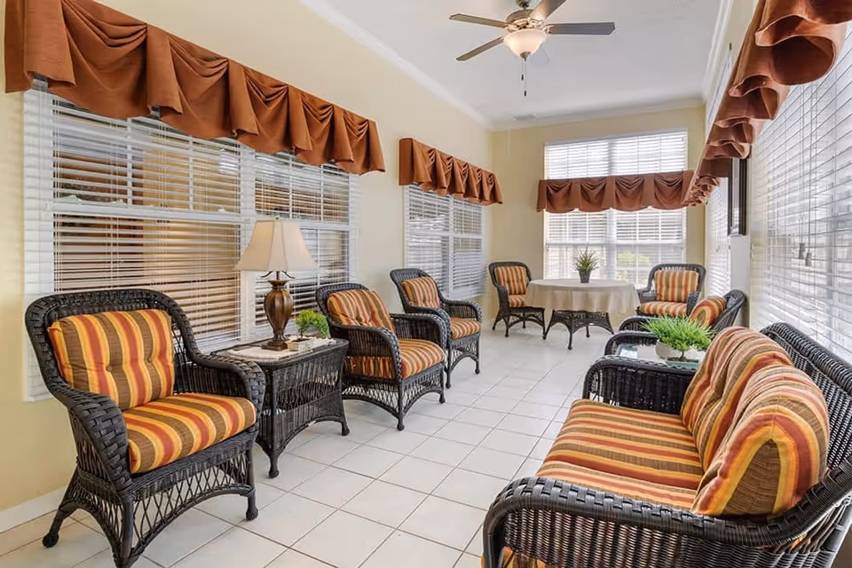 A bright sunroom with multiple wicker chairs and a wicker loveseat, all with orange and yellow striped cushions. There are two small wicker side tables with plants and a lamp, and a round table with four chairs in the background. The room has large windows with white blinds and brown valances, a ceiling fan with a light, and white tiled flooring.