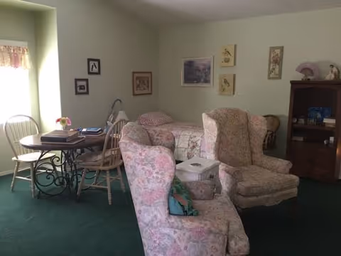 Cozy living area with floral upholstered armchairs, a small round table by a window, and a bed against a pale green wall.