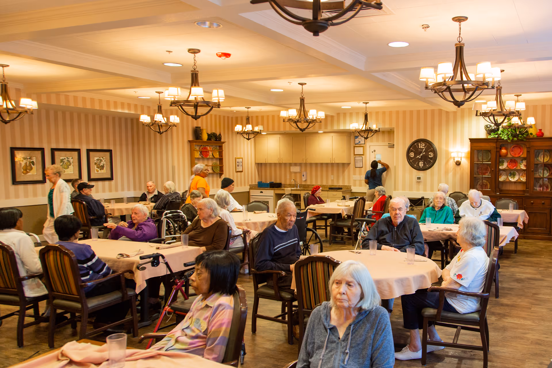 A dining room in an assisted living facility with elderly residents seated at round tables covered with light pink tablecloths. The room has warm lighting from chandeliers and wall sconces, striped wallpaper, framed artwork, and a large clock on the wall. Some residents are engaged in conversation while others sit quietly. A kitchen area is visible in the background.