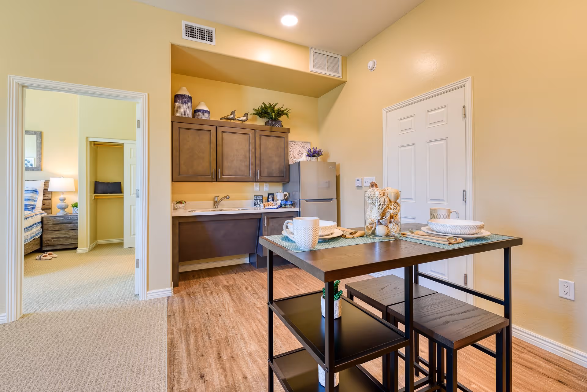 A small kitchen area with wooden cabinets, a sink, and a refrigerator. In front of the kitchen is a dining table set with plates, cups, and decorative items. To the left, there is an open doorway leading to a bedroom with a bed, nightstand, and lamp. The room has light yellow walls and wood flooring in the kitchen area, with carpet in the bedroom.