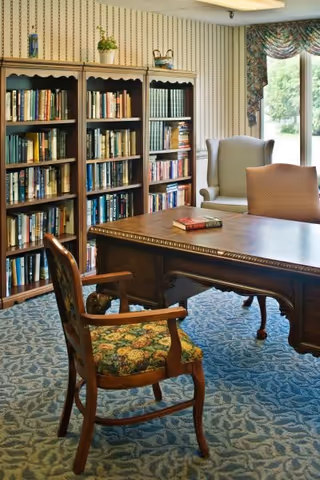 A cozy interior room featuring a wooden table with two books on it, surrounded by three chairs including one with floral upholstery. Behind the table are three tall bookshelves filled with books. The room has patterned wallpaper, a blue carpet, and a large window with floral curtains letting in natural light.