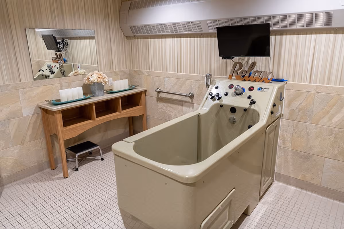 A therapeutic bathing room with a beige walk-in bathtub equipped with multiple control knobs and a mounted TV above it. A wooden table with three compartments holds a flower arrangement and candles. The walls are tiled halfway up with beige tiles and striped wallpaper above. A mirror and a small step stool are also visible.