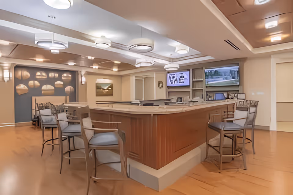 Interior view of a senior living facility common area with a large wooden bar counter surrounded by high chairs. The room has warm lighting with ceiling fixtures, two wall-mounted televisions, and decorative wall art. The floor is wooden, and the space appears clean and inviting.