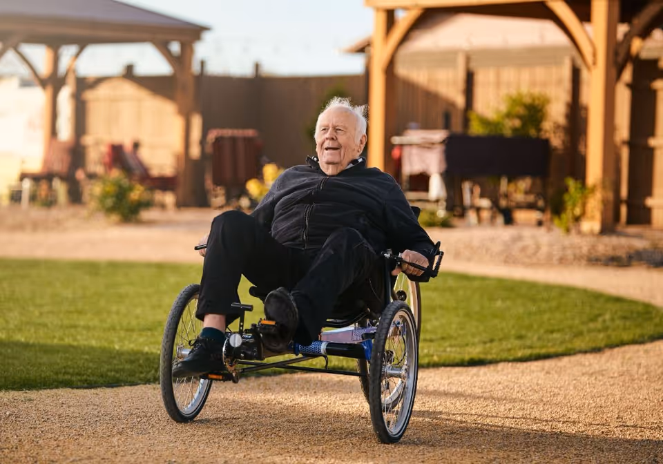 An elderly man riding a three-wheeled recumbent bicycle on a gravel path in an outdoor garden area with wooden pergolas and green grass.